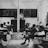 A classroom setting with several students seated at individual desks, all facing a teacher standing near a blackboard. The room has simple furniture and chalkboards with illustrations, including a map of Africa and drawings of a cow and tree.